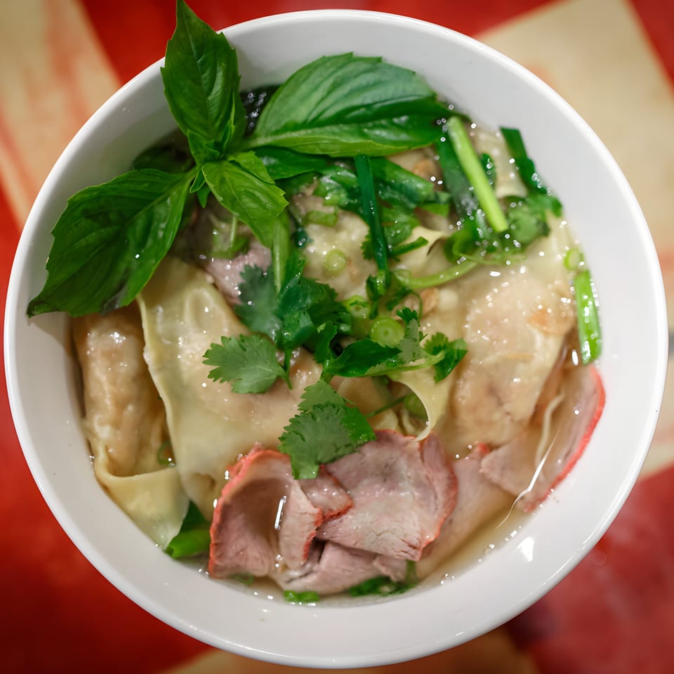 Overhead view of Vietnamese wonton soup with sliced pork, basil, and cilantro in a white bowl.